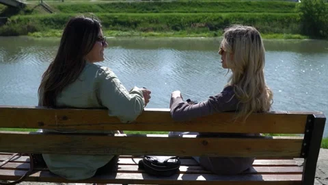 Two young ladies having a chat while sitting on a bench near a river. Stock Footage 304174683