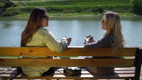 Two young ladies having a chat while sitting on a bench near a river. Video stock 304175581