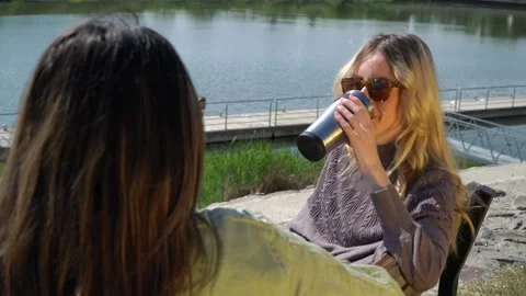 Two young ladies having a chat while sitting on a bench near a river. Video stock 304176230