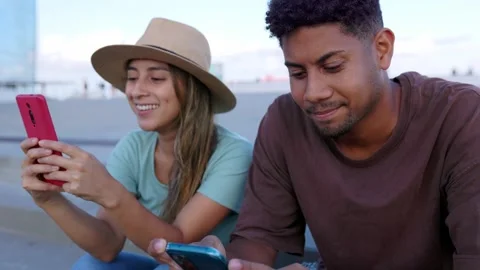 Two young latin friends using cell phones sitting together outdoors. Stock Footage 250837844