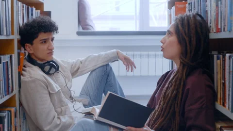 Two Young Learners Lost in Conversation with Open Books Between Library Shelves Vidéo 335131369