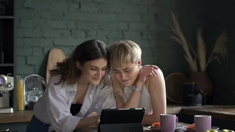 Two young lesbian women using tablet while sitting at table in home kitchen Stock Footage 128288759