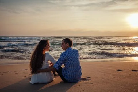 Two young lovers sitting on a beach and looking to each other on sunset 스톡 사진