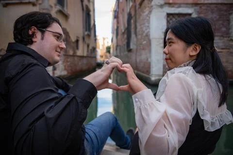 Two young lovers sitting in front of the canal draw a heart with their finger Stock Photos