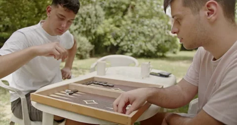 Two young men are engaged in a game of backgammon in a peaceful garden setting Stock Footage 306807707