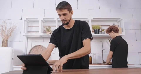 Two young men are using a digital tablet and laptop in the kitchen Stock Footage 247472181