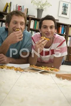 Two young men lying on floor, eating pizza Stock Photos