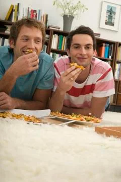 Two young men lying on floor, eating pizza Stock Photos