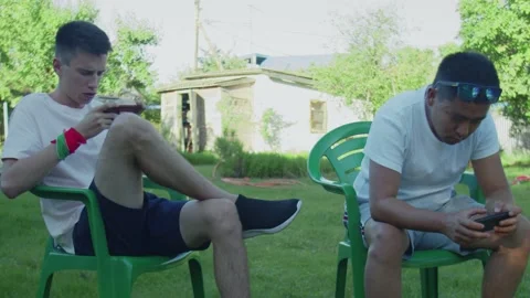 Two young men plays together on phones in multiplayer, sitting on chairs on Stock Footage 143488193