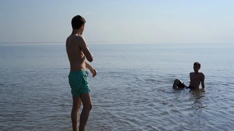 Two young men in shallow water, one walks out of the water 库存影片 90805912