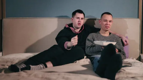 Two young men sitting on a bed, sharing a bowl of chips while talking and Stock Footage 304817689