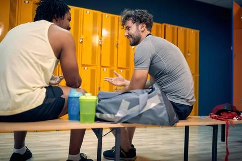 Two young men sitting on bench in dressing room, having a fun conversation .. Stock Photos