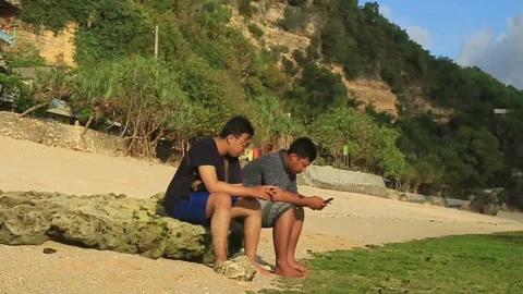 Two young men sitting on a rock playing with their cell phones Video stock 275344410