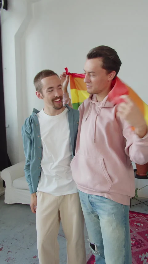 Two young men stand together, covered with a lgbt flag, and look at the camera Stock Footage 173902092