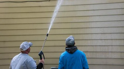 Two young men standing by the side of a yellow house. One is power washing th Stock Photos