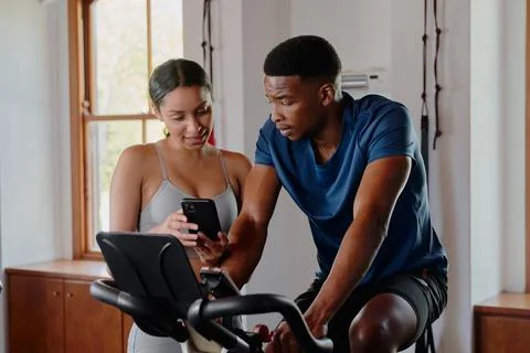 Two young multiracial friends using mobile phone on exercise bike at the gym Stock Photos