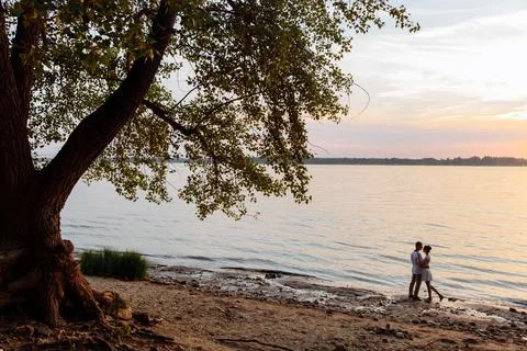 Two young people hug under an old tree by the river. Stock Photos
