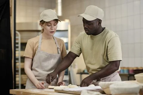 Two young people working in bakery kitchen with young woman instructing trainee Stock Photos