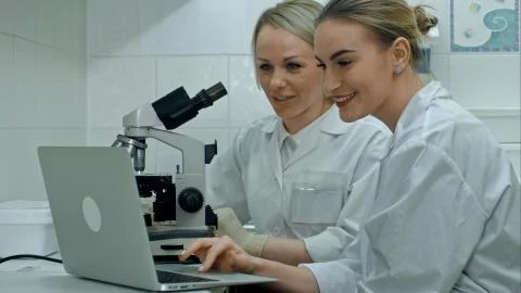 Two young positive scientists working in laboratory using microscope and laptop Stock Photos