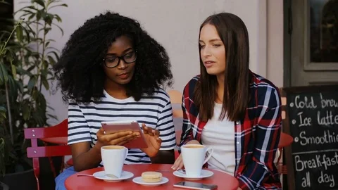 Two young pretty friends sitting at table with coffee in cafe and viewing photos Stock Footage 81834614