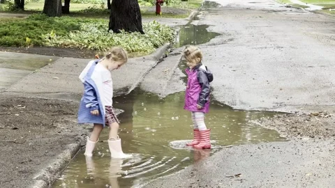 Two young sisters in bright raincoats wade in a puddle Video stock 233740286