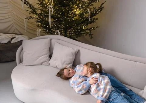 Two young sisters cuddle together on a light gray sofa, enjoying the warm glo Stock Photos