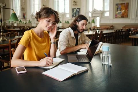 Two young students making study notes with laptop in library of university Stock Photos