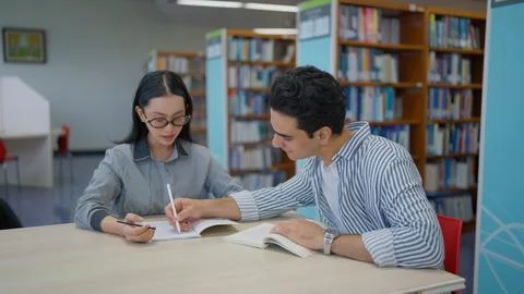 Two young students sit at a library table studying together with books and pens Stock Photos