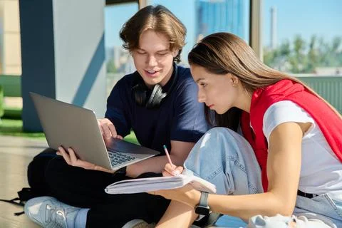 Two young students studying with laptop and notebook sitting on floor near big Stock Photos