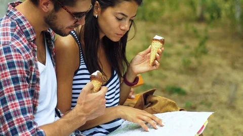 Two young tourist sitting on the bench, holding map on hands and eating tasty Stock Footage 70373329