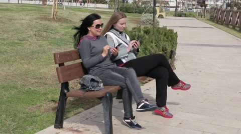 Two young woman using a smart phone, outdoors on sitting on the wooden bench in Stock Footage 62112059