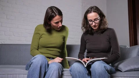 Two young women on a gray sofa sharing a bookone reading aloud while the other Stock Footage 321262389