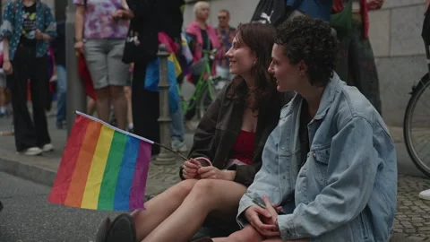 Two young women hold a small rainbow flag while sitting together on a curb Stock Footage 346239962