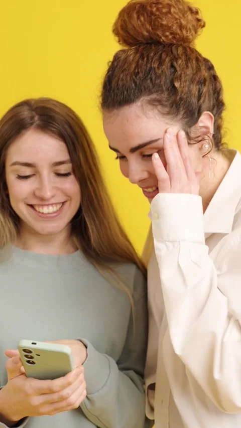 Two young women sharing smartphone content on yellow background Video stock 308653640