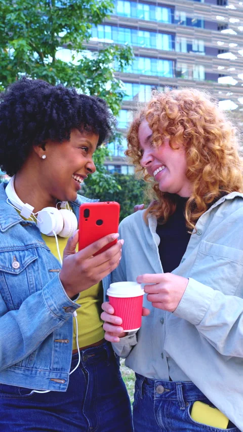 Two young women smiling while using smartphones outdoors in casual clothing Stock Footage 314887576
