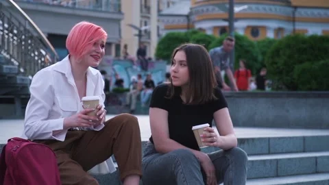 Two young women talking and laughing sitting on the granite stairs and drinking Stock Footage 141498677