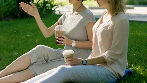 Two young women talking, drinking hot coffee and cold juice in a summer park. 4K Stock Footage 87725439
