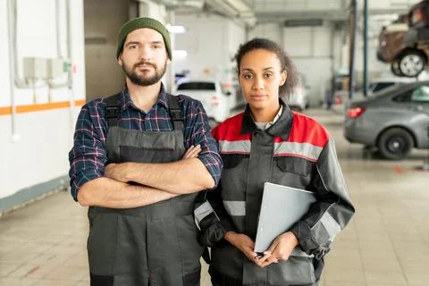 Two young workers of car repair center standing against automobiles in workshop Stock Photos