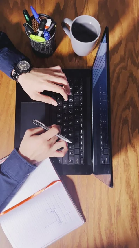 Typing on a laptop and holding a pen at a wooden desk Stock Footage 345149643