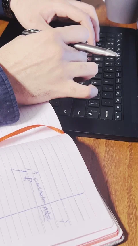 Typing on a laptop keyboard and holding a pen with an open notebook Stock Footage 345150953