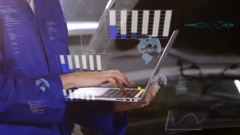 Typing tech in blue coveralls using silver laptop at bay beside car hood showing 库存影片 333962652