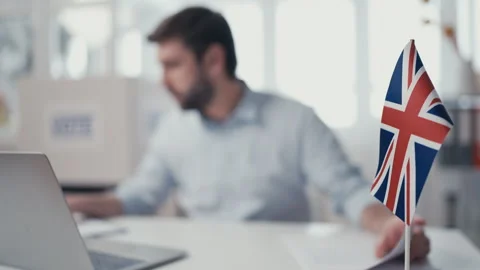 UK embassy employee processing documents, typing on laptop, British flag on desk Stock Footage 240835778