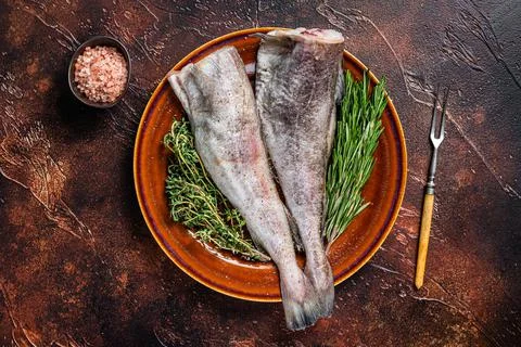 Uncooked Raw cod or codfish fish in rustic plate with herbs and fork. Dark Stock Photos