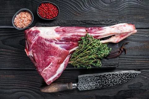 Uncooked Raw lamb mutton leg with herbs on butcher cutting board. Black wooden Stock Photos