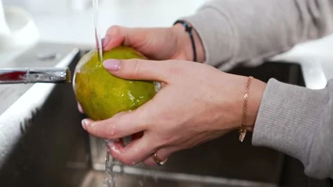 Under a warm stream of water from the tap, a woman washes a mango fruit for Stock Footage 197051956