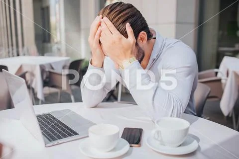 Unhappy and sad young man sits at table in restaurant. He closed his face with Stock Photos