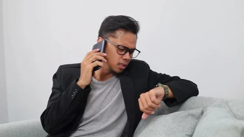 Unhappy asian man checking time and calling a friend while sitting on the sofa Stock Footage 336510032