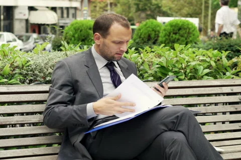 Unhappy businessman comparing data on smartphone and documents on bench NTSC Stock Footage 43337685