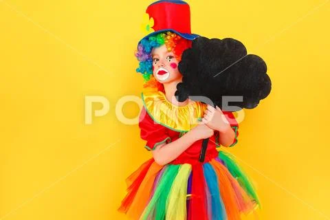 Unhappy girl in clown costume and hat holding black table Stock Photos