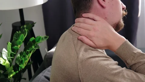 Unhappy guy sitting on couch holding hand on neck, suffering from neck pain Stock Footage 225229772